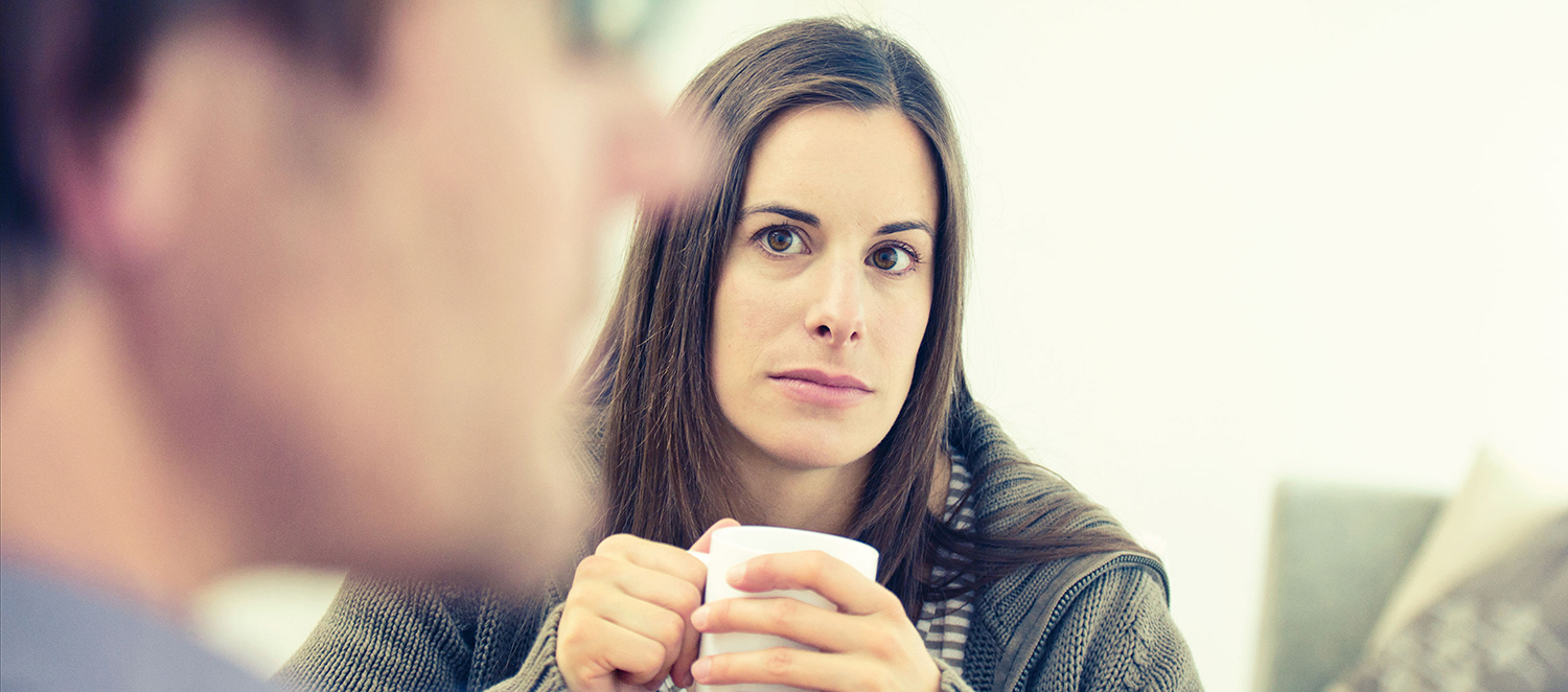 Eine Frau schaut nachdenklich, eine Tasse umklammernd, ihren Partner an, der sich schemenhaft im Vordergrund befindet.