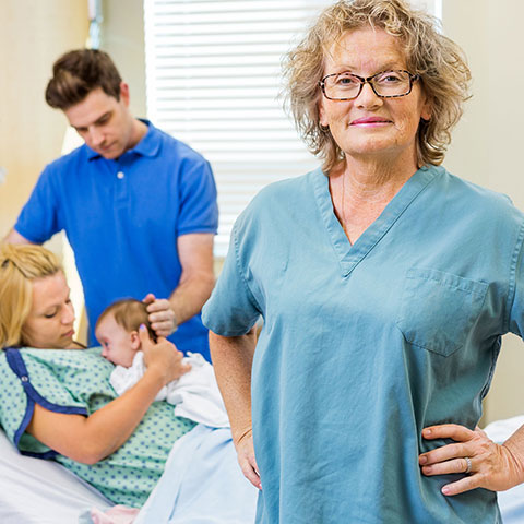 Im Krankenhaus-Zimmer: Lächelnde Hebamme im Vordergrund, im Hintergrund Paar mit Neugeborenem.