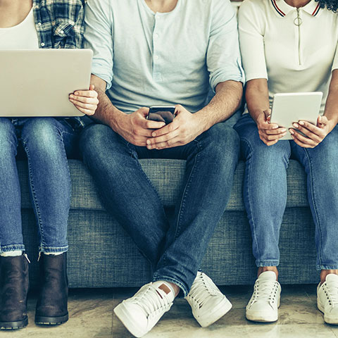 Fünf Erwachsene sitzen nebeneinander auf dem Sofa und bedienen Smartphones, Tablet und Laptop. 